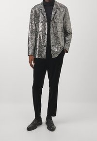 Blazer argento con paillettes e collo a scialle, tasca frontale e dolcevita nera. Abbinato a pantaloni in velluto nero e scarpe eleganti nere.