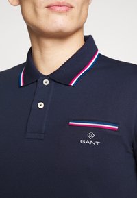 Granatowa koszulka polo z prążkowanym kołnierzykiem w czerwono-niebiesko-białe paski. Dwa guziki na plastrze oraz mała kieszonka na piersi z akcentem w paski.