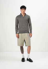 Maglione grigio con zip e maniche lunghe, abbinato a pantaloni corti beige. Il modello indossa mocassini neri e calzini bianchi; è visibile un tatuaggio su una gamba.