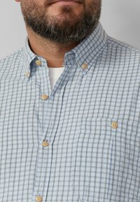 Lichtblauwe en witte geruite shirt met knoopsluiting, houten knopen en een borstzak. De stof lijkt zacht en gestructureerd.