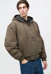 Smešani smeđi dukserica na zatvaranje sa velikim kapuljačom, prednjim džepovima i rebrastim manžetama. Nosena preko crne košulje s karo uzorkom, koja ima aplikaciju s logotipom.