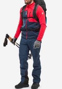 Rote und marineblaue Softshelljacke, graue Handschuhe, marineblaue Hose mit schwarzen Akzenten und ein schwarzer Rucksack. Ausgestattet mit Wanderstöcken.