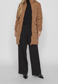 Veste longue matelassée marron avec un col haut, une fermeture éclair et des poches latérales, associée à un pantalon noir à jambes larges et des chaussures noires à bouts pointus.