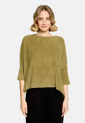 Femme aux cheveux blonds courts portant un pull ample et duveteux vert olive et un pantalon noir, debout devant un fond clair uni.