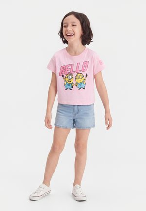 Lachend kind staat met een roze Minions "Bello" T-shirt, spijkershorts en witte sneakers, voor een effen witte achtergrond.