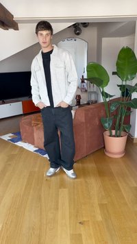 Helehall hall triiksärk musta T-särgi peal, kombineeritud tumedate teksade ja hõbedaste tossudega. Siseruumides, taustal taimed ja mööbel.