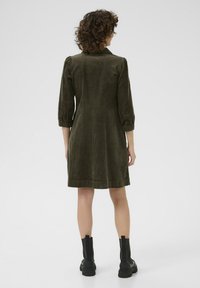 Abito in corduroy khaki con maniche tre quarti, corpino aderente, gonna svasata e tessuto testurizzato; indossato con stivaletti neri alla caviglia.