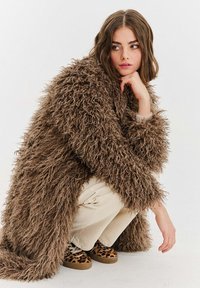 Hnědý načechraný dlouhý kabát s texturovaným materiálem, krémové kalhoty a tenisky s leopardím vzorem. Modelka je v podřepu a opírá si ruku o bradu.