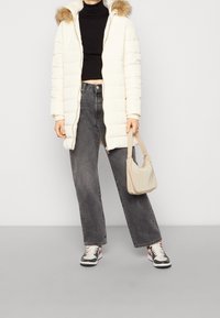 Abrigo de invierno acolchado blanco con capucha forrada de pelo, jersey negro de cuello alto acanalado, vaqueros grises de pierna ancha y un bolso de hombro color beige claro. Se presentan unas zapatillas negras.