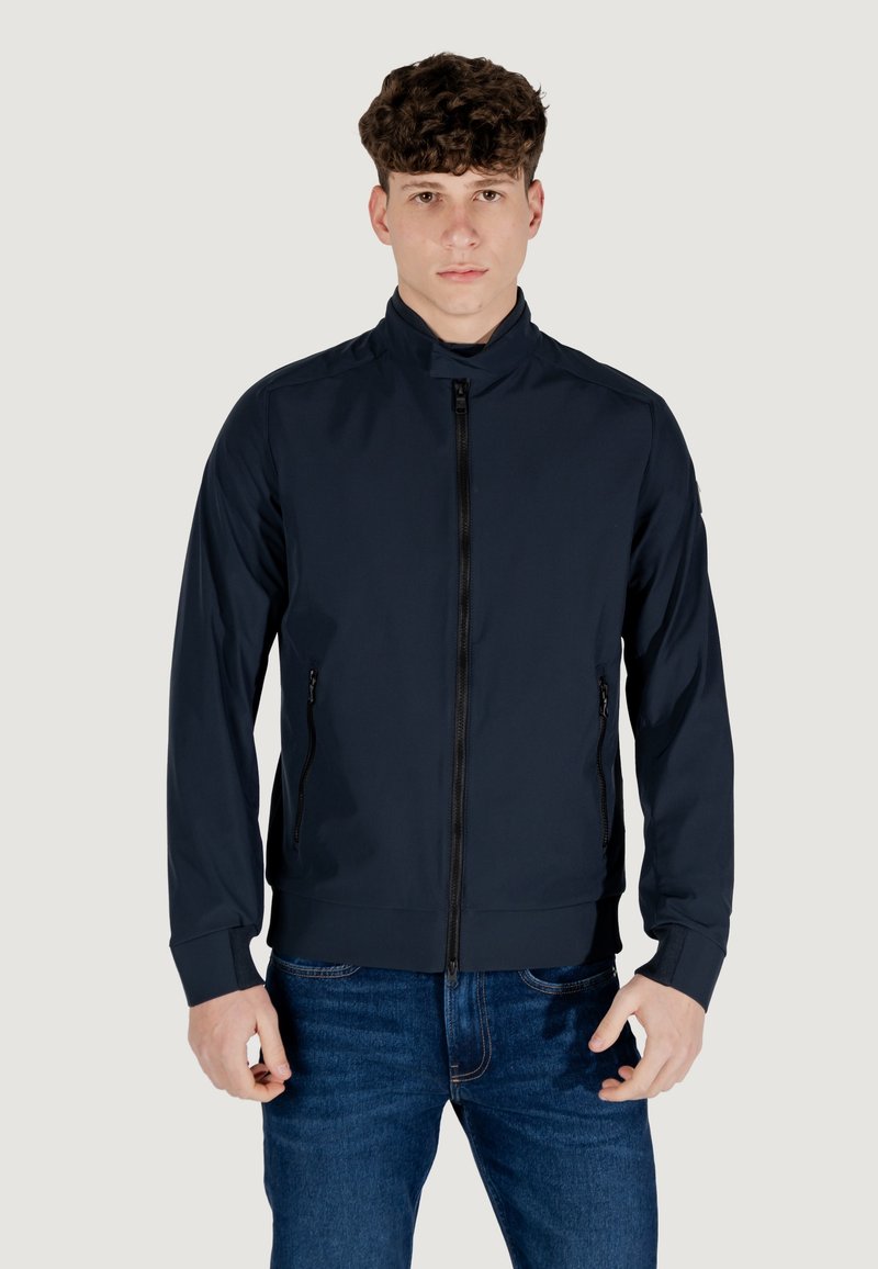 Giacca zip a collo alto di colore blu navy, con due tasche laterali e un design aderente. Realizzata in materiale liscio e leggero con polsini a coste.