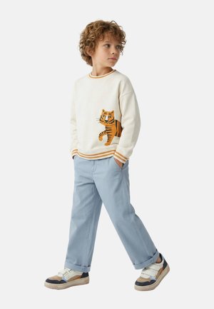 Jeune garçon aux cheveux bouclés portant un sweat-shirt blanc avec un motif de tigre, un pantalon bleu clair et des baskets multicolores, debout les mains dans les poches.