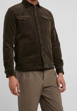 Man draagt een donkerbruine corduroy jas met knopen en klepzakken en beige broek tegen een effen achtergrond.