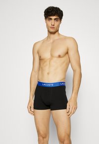 Schwarze Boxershorts mit einem blauen Lacoste-Logo-Bund. Der Stoff wirkt glatt und dehnbar, was eine enge Passform bietet.