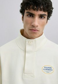 Suéter de color crema con un cuello alto, que presenta un destacado parche de logo en amarillo y azul y un cierre de botón en el cuello.