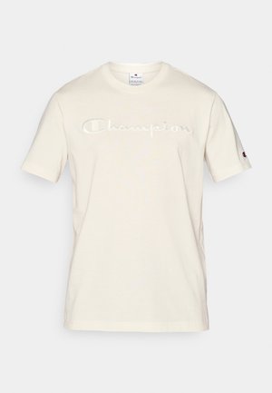Majica od pamučne tkanine krem boje s kratkim rukavima, na kojoj je izvezen "Champion" logo u malo tamnijoj nijansi preko prsnog dijela.