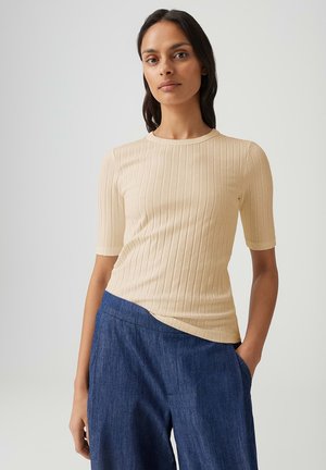 Vrouw met lang donker haar, gekleed in een beige geribd kort mouwen topje en blauwe wijde broek, staand met één hand in de zak.