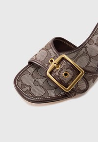 Sandale slide en tissu marron avec un design en maille, une grande boucle dorée et des détails de couture sur les brides et la semelle.