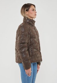 Giacca a vento marrone con colletto alto e motivo geometrico. Caratterizzata da una chiusura con zip e una texture trapuntata, abbinata a jeans di un denim chiaro.