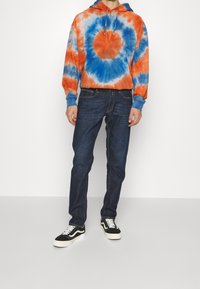 Sudadera tie-dye en naranja y azul, material de algodón, capucha con cordones, bolsillo frontal, combinada con jeans azul oscuro y zapatillas negras.