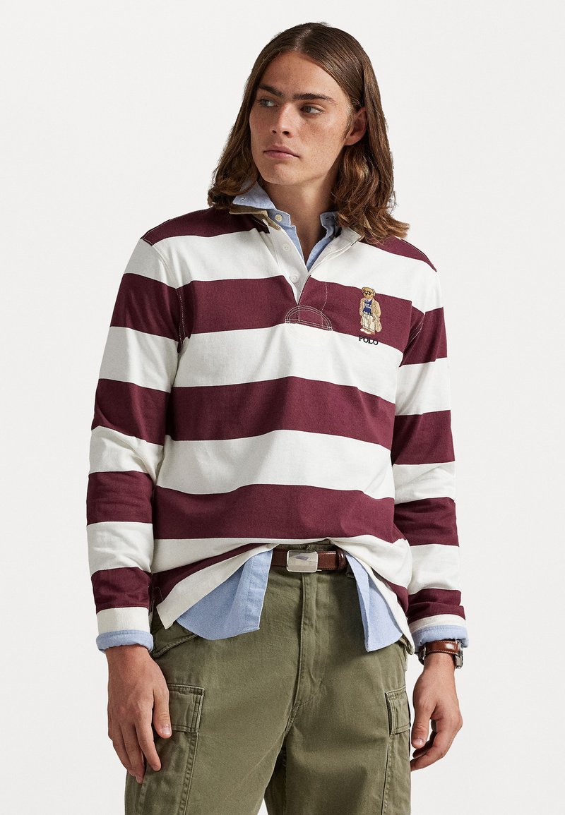 Tânăr cu părul până la umeri, îmbrăcat cu un tricou de rugby alb cu dungi maronii peste o cămașă albastră și pantaloni cargo verzi.