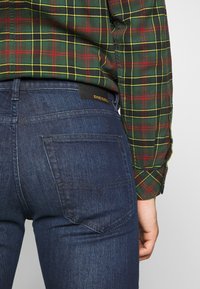 Mörkblå denimjeans med en slät textur, som har en svart "DIESEL"-logotyp och enkla bakfickor. Bärs med en grön rutig skjorta.