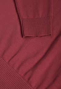 Gros plan sur un poignet et une ceinture côtelés d'un pull en maille à manches longues de couleur rouge foncé.