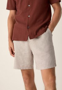 Hombre con pantalones cortos de lino beiges con bolsillos y una camisa de botones marrón de manga corta con textura, de pie con una mano en el bolsillo.