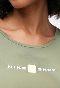 Κοντινό πλάνο ενός ατόμου που φοράει πράσινη μπλούζα Nike Shox, με ορατά τα κόκαλα του στήθους και το λογότυπο στο κέντρο του στήθους.