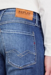 Blå denimjeans med en brun läderlapp som har "REPLAY BLUE JEANS," distinkta sömmar på fickorna och subtila texturdetaljer.