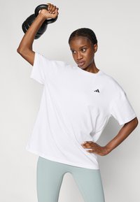 T-shirt en coton blanc à manches courtes avec un petit logo noir, associé à des leggings vert clair. La personne tient un kettlebell noir au-dessus de sa tête.