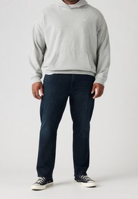 Šedá bavlněná mikina s kapucí a přední kapsou, s ribovanými manžetami, kombinovaná s tmavě modrými džíny a černými teniskami s bílými akcenty.