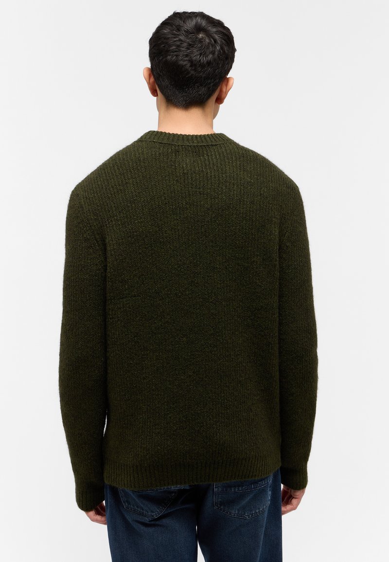 Hombre con pelo corto y oscuro, vistiendo un suéter de punto verde oliva y jeans azules, de pie de espaldas contra un fondo blanco.