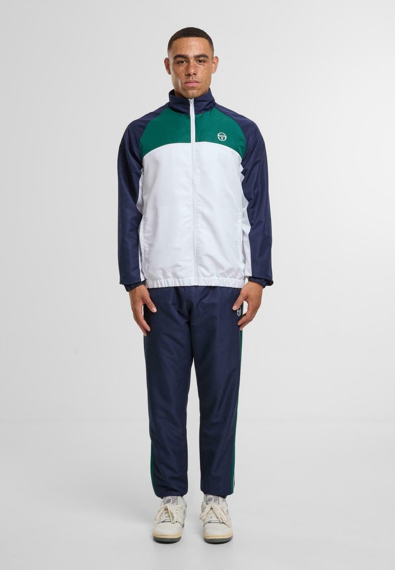 Tuta sportivo con base blu navy, parte superiore bianca e dettagli verdi. Presenta una chiusura con zip e polsini elastici. Indossato con scarpe da ginnastica bianche.