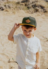 Cappello verde con visiera gialla e logo, camicia button-up chiara, sfondo sabbioso, bambino che solleva il cappello, maniche corte.