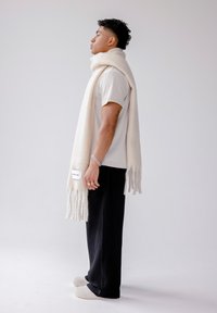 Witte, zachte sjaal met franjes aan de uiteinden, gedragen door een model. De sjaal heeft een label en is gecombineerd met zwarte geribbelde broek en beige schoeisel.