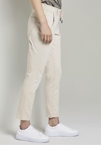 Helle beigen Hosen mit schmaler Passform, seitlichen Taschen und einem Kordelzugbund, kombiniert mit weißen Sneakers. Glattes Material mit minimaler Textur.