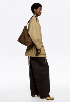 Chaqueta beige de gran tamaño, pantalones marrones sueltos, bolso marrón y zapatos texturizados. Paleta de colores tierra con diseño minimalista y detalles funcionales.