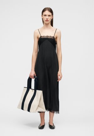 Γυναίκα που φοράει μαύρο φόρεμα με λεπτομέρειες από δαντέλα, κρατώντας μια μεγάλη λευκή τσάντα ώμου με μαύρα λουριά, στέκεται μπροστά σε απλό φόντο.