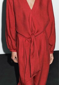 Rød linned jumpsuit med V-hals og lange ærmer, der har en knyttet taljedeatil. Stoflet har en tekstureret, let krøllet overflade.