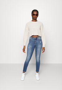 Cremefarvet cropped striktrøje, blå skinny jeans med slidte detaljer og hvide sneakers. Tilbehøret inkluderer brune solbriller.