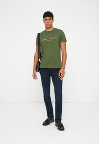 Grøn bomulds T-shirt med et hvidt logo. Sammen med mørkeblå jeans og sorte sko, og bærer en crossover taske.