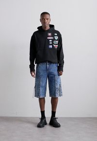 Black hooded sweatshirt with patches, paired with blue denim shorts featuring pockets and fringe details. Black socks and shoes complete the look.