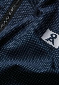 Marineblauer Stoff mit weißem Gittermuster, Nahaufnahme eines schwarzen Reißverschlusses und eines teilweisen weißen Etiketts mit schwarzem stilisiertem "RA"-Schriftzug.