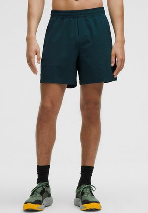 Ciemnozielone sportowe szorty z elastycznym pasem, sięgające kolan, o gładkiej fakturze. Noszone z zielonymi butami i czarnymi skarpetkami.