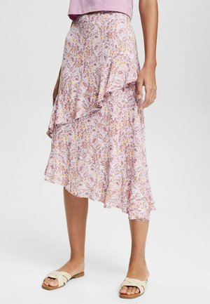 Vrouw draagt een roze midi rok met bloemenprint en asymmetrische ruches, beige platte sandalen, staand op een effen lichte achtergrond.