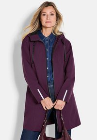 Paarse waterdichte jas met een capuchon, voorzien van zwarte accenten, witte reflecterende details op de mouwen en een rits voorziening over een denim shirt.