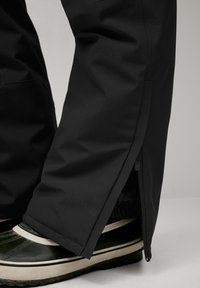 Black waterproof pants with a zipper detail at the ankle, paired with green and black high-top shoes featuring a white sole.
