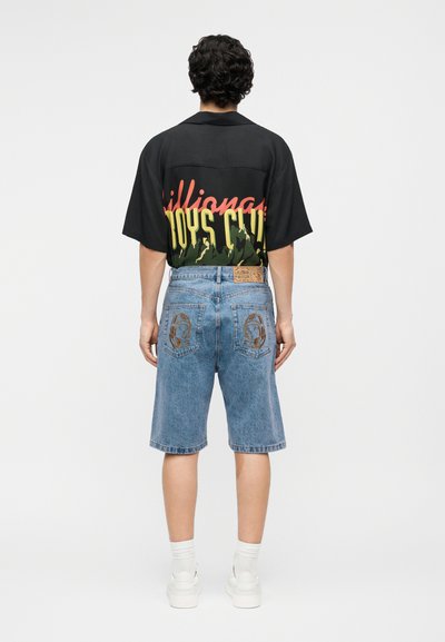 Férfi háttal, fekete mintás rövid ujjú pólót és középhosszú kék farmer rövidnadrágot visel, az űrhajós motívumok a hátsó zsebeknél láthatók.