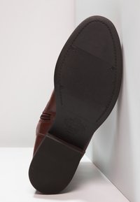 Botín de cuero marrón con suela de goma negra. Presenta cremallera lateral, costuras decorativas y una puntera redondeada con patrón de agarre texturizado.