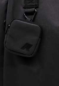 Borsa in tessuto nero con una piccola tasca con zip, caratterizzata da una superficie testurizzata e un logo geometrico, attaccata con una robusta cinghia e hardware.